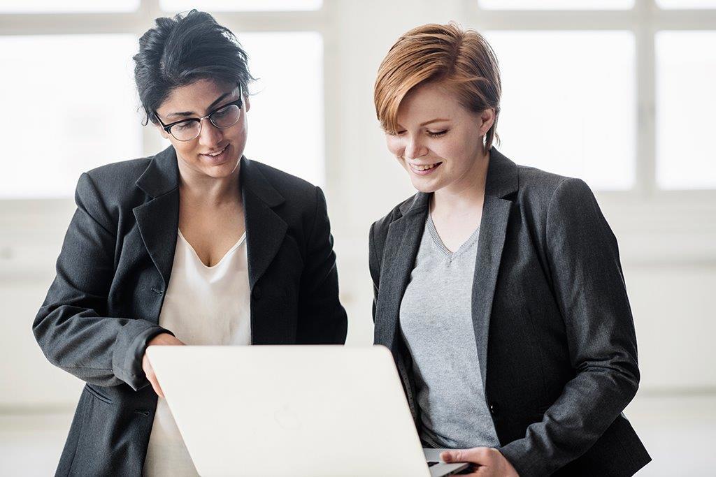 Two professional women in business attire collaborating on a laptop, engaged in discussion in a bright office setting.