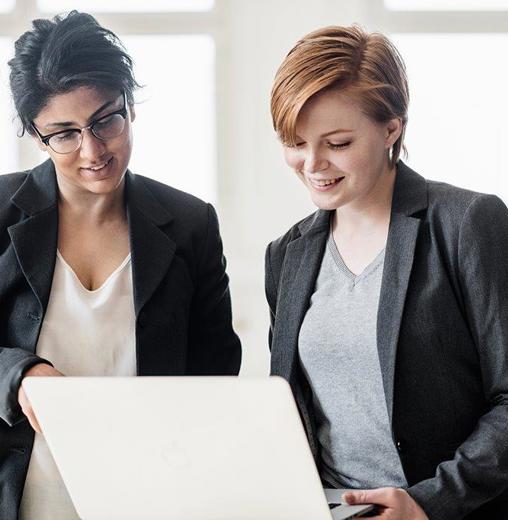 Two professional women in business attire collaborating on a laptop, engaged in discussion in a bright office setting.