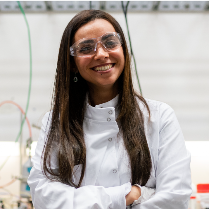 A smiling female scientist wearing protective glasses and a white lab coat stands in a laboratory. She appears confident and focused, with scientific equipment visible in the background.