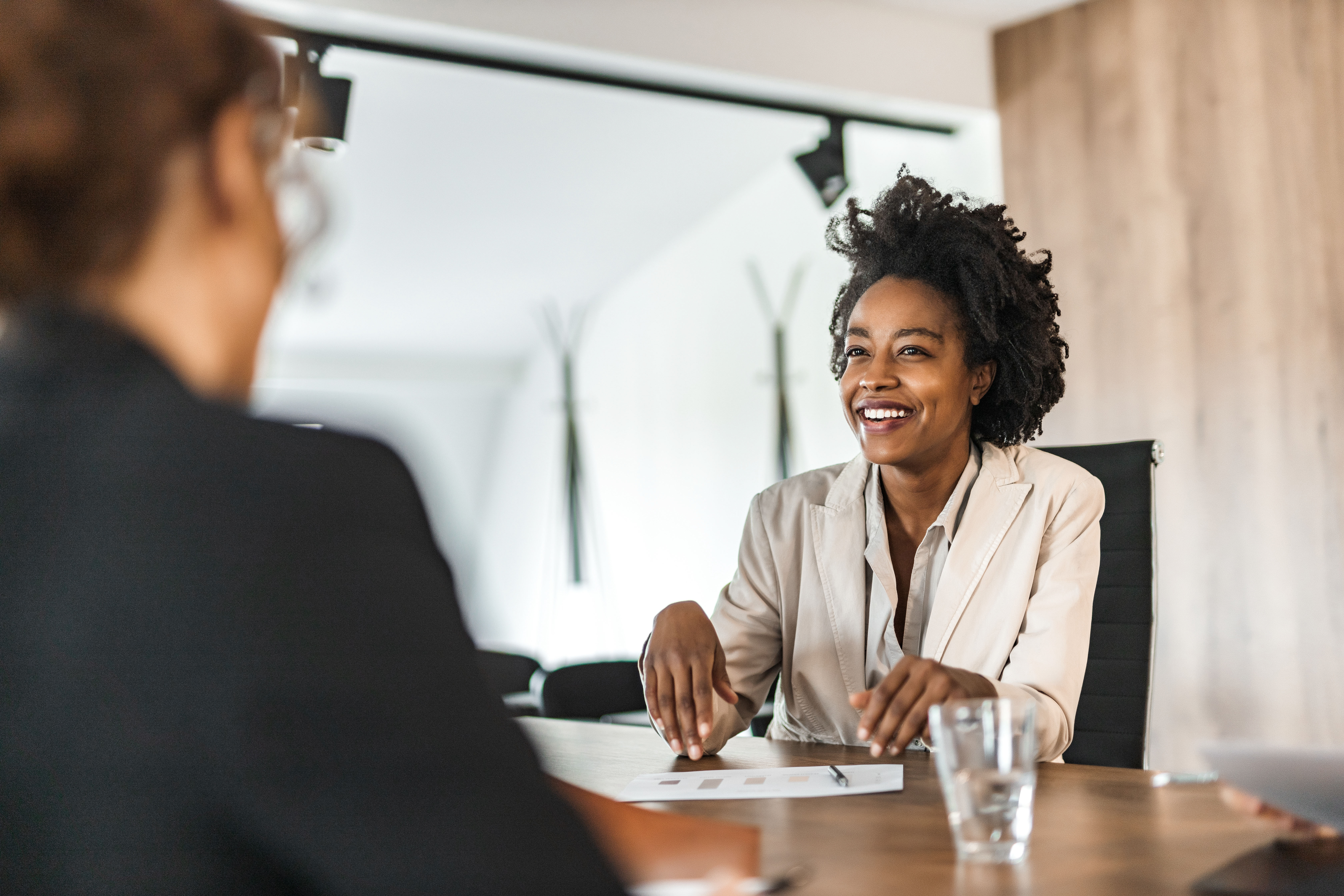 A confident and smiling woman in a beige blazer is engaged in a conversation during a professional meeting. She is seated at a table with documents and a glass of water, conveying enthusiasm and positivity. The setting is a modern office space, suggesting a job interview, business negotiation, or networking discussion.