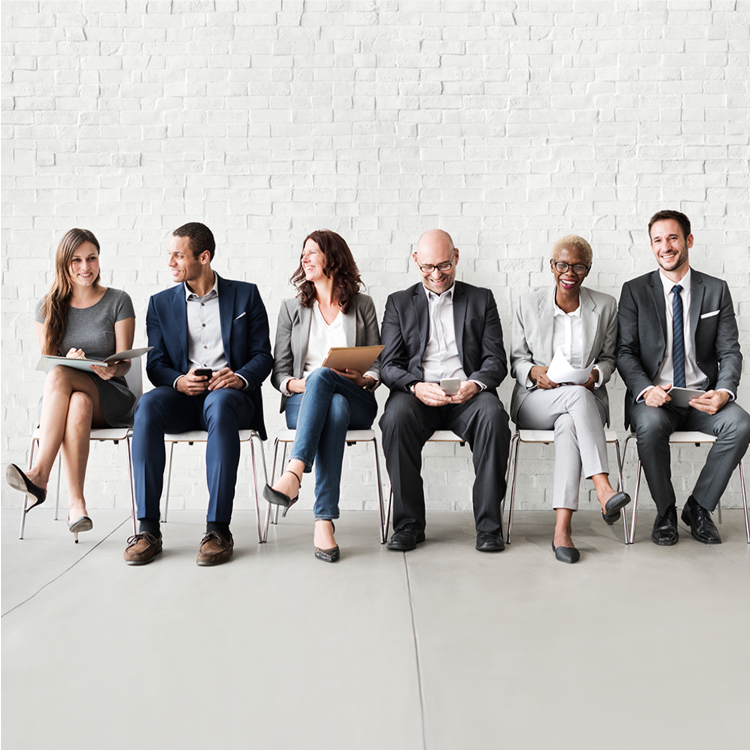 A diverse group of six professionals sitting in a row against a white brick wall, engaged in conversation and smiling, representing an inclusive and dynamic workplace environment.
